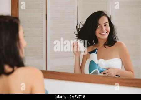 Beautiful happy young woman in bath towel drying hair and looking into the mirror in bathroom