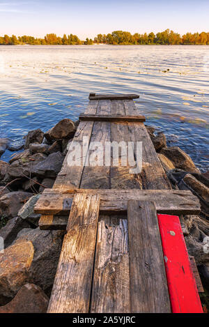 A wooden pontoon with a red paint plank  over big stones close to the blue Dnieper river is waiting for the fisherman. Stock Photo