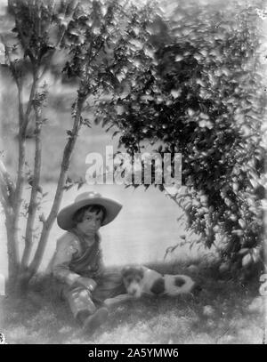 Photograph of a Boy with a dog, a study made at Oceanside. Black and White. The boy is wearing overalls and a hat, sat next to a tree on the ground. The dog sits next to him in the shadow of shrubbery. In the background is a pond. Circa 1904 Gertrude Käsebier, 1852-1934, photographer. Stock Photo