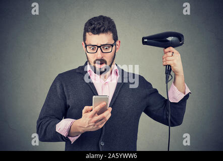 Shocked beard man checking his phone holding a hair dryer Stock Photo
