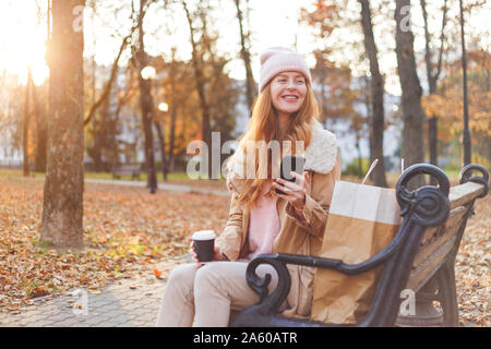 Redhead girl in warm clothes sitting with smartphone on wooden bench in autumn park. Stock Photo