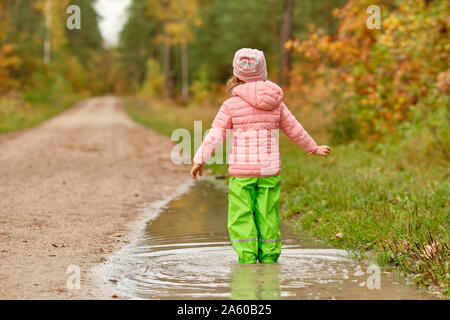 Rear view of a caucasian child girl with the arms strechted out in green waterproof pants and rubber boots standing in a huge rain puddle alongside a Stock Photo