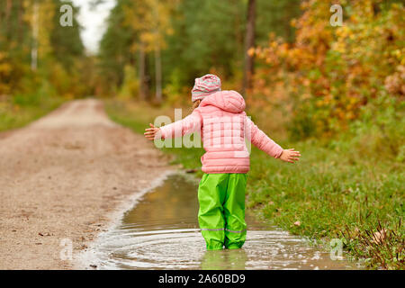 Rear view of a caucasian child girl with the arms strechted out in green waterproof pants and rubber boots standing in a huge rain puddle alongside a Stock Photo