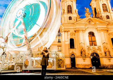 Woman standing in front of a colorful ferris wheel drawing a light heart in the air at winter time with a church in the background Stock Photo