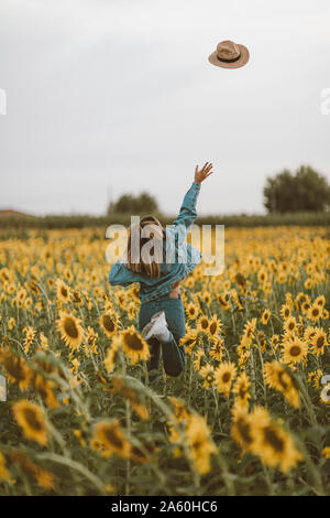 Rear View of young woman with blue denim jacket throwing a hat in a field of sunflowers Stock Photo
