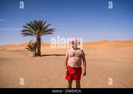 Overweight man with swimming shorts and hat standing in the desert of Morocco Stock Photo