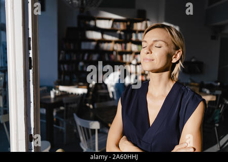 Businesswoman enjoying the sun, with eyes closed, standing in door of a coffee shop Stock Photo