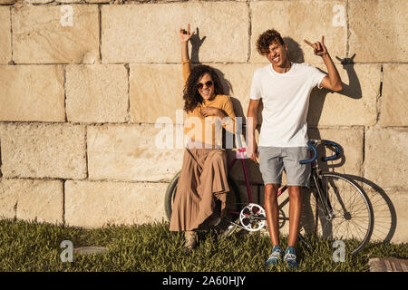 Young couple with bicycle, leaning on stone wall, making hand signs Stock Photo