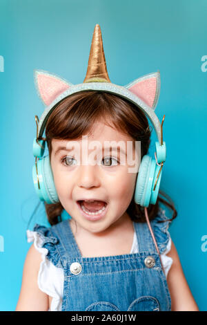 Portrait of cute little girl listening music and singing with unicorn shaped earphones on blue background Stock Photo