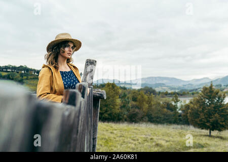 Young curly haired woman wearing a hat, yellow coat and blue t-shirt looking the mountain landscape Stock Photo