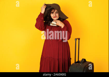 Attractive south asian traveler woman in deep red gown dress posed at studio on yellow background with suitcase and credit cards. Stock Photo