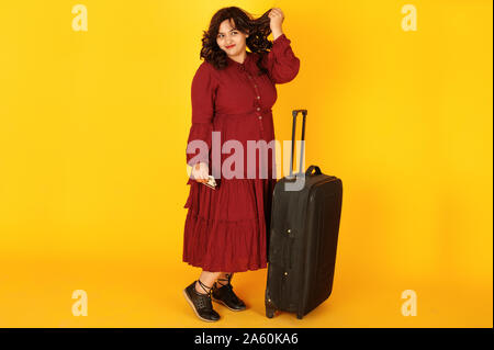 Attractive south asian traveler woman in deep red gown dress posed at studio on yellow background with suitcase and credit cards. Stock Photo