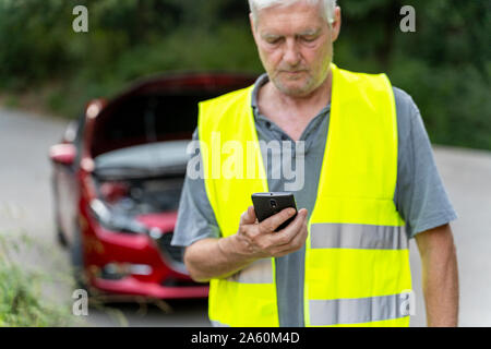 Senior man standing at his broken car wearing a safety vest and using his smartphone Stock Photo