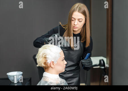 Professional hairdresser is dyeing female hair in a beauty salon. Woman hairdresser dyes hair in white color at beauty salon. Stock Photo