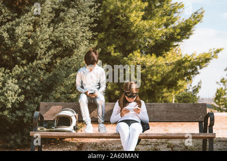 Two kids in astronaut and superhero costumes using mobile phone on park bench Stock Photo