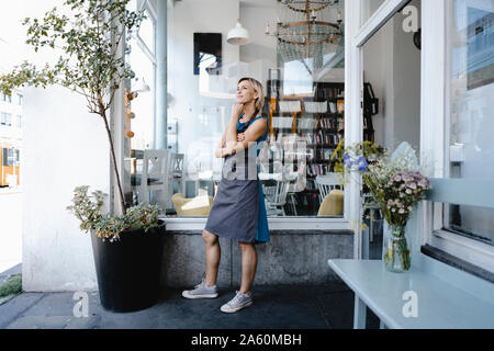 Woman with apron standing in front of coffee shop, thinking Stock Photo