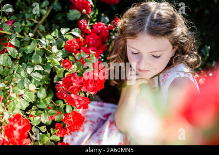 Portrait of girl with eyes closed sitting beside red rosebush Stock Photo