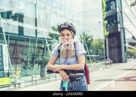Portrait of happy young woman  wearing cycling helmet leaning on handlebar of E-Scooter Stock Photo