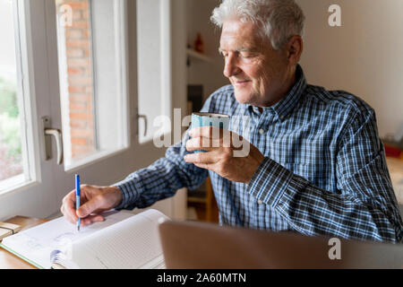Senior man sitting at home in front of laptop making notes Stock Photo
