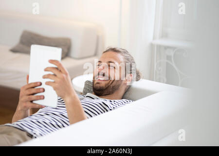 Smiling young man lying on couch at home using tablet Stock Photo