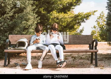 Two kids in astronaut and superhero costumes using mobile phone on park bench Stock Photo