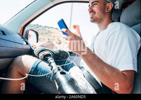 Smiling young man with leg prosthesis sitting in camper van using smartphone Stock Photo