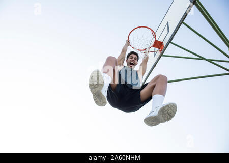 Young man screaming while hanging on basketball hoop and looking at camera Stock Photo