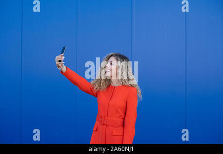 https://l450v.alamy.com/450v/2a60nj1/smiling-young-woman-wearing-red-dress-taking-selfie-in-front-of-blue-background-2a60nj1.jpg