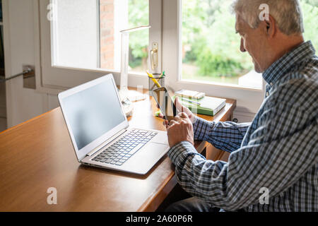 Senior man sitting in front of laptop using smartphone at home Stock Photo