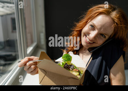 Portrait of happy redheaded businesswoman eating healthy takeaway food and talking on the phone Stock Photo