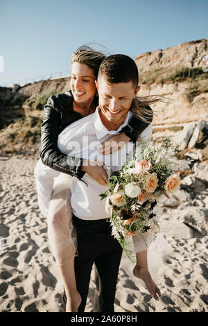 Happy groom carrying bride piggyback on the beach Stock Photo