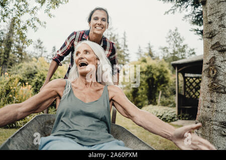 Daughter pushing mother, sitting in push cart, through the garden Stock Photo