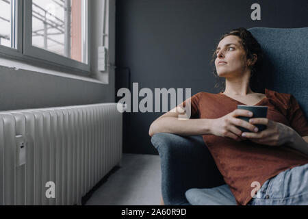 Young woman with cup of coffee on lounge chair looking out of window Stock Photo
