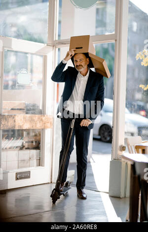 Businessman with cardboard box on his head, riding e-scooter in a coffee shop Stock Photo