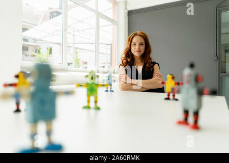 Portrait of redheaded businesswoman in a loft with miniature robots on desk Stock Photo