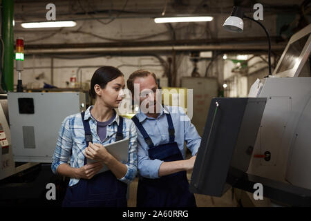 Bearded young engineer working on machine and showing something to young woman with tablet pc in the plant Stock Photo