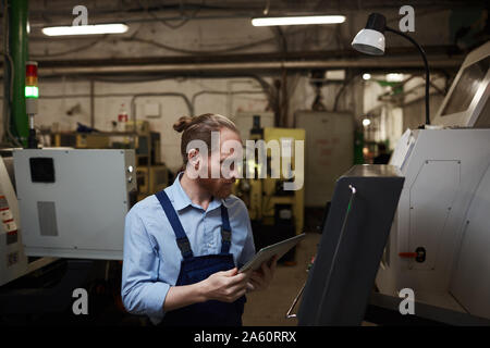 Serious manual worker in overalls using tablet pc and controlling machine remotely in the factory Stock Photo