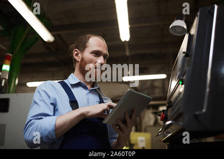 Young bearded engineer typing on digital tablet and looking at monitor of machine in the factory Stock Photo