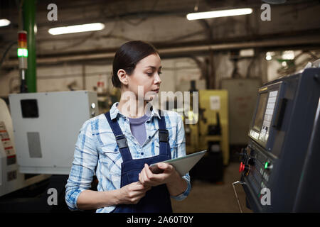 Serious young engineer in work wear holding digital tablet and looking at machine she controlling the work Stock Photo