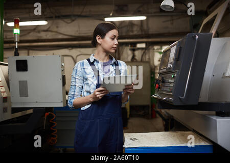 Serious technician in overalls controlling the work of machine using her digital tablet in the plant Stock Photo