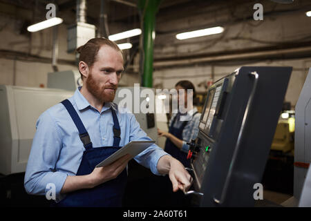 Serious bearded engineer controlling the work of machine with tablet pc with his colleague working in the background Stock Photo