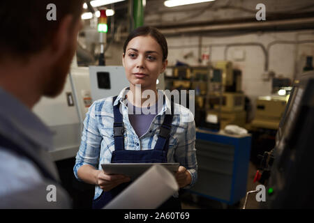 Young businesswoman in special work wear holding digital tablet and talking to the manual worker during his work in the plant Stock Photo