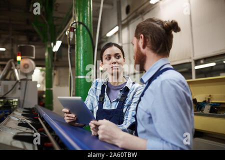 Young female worker holding digital tablet and talking to her colleague while they standing near the machine in the plant Stock Photo