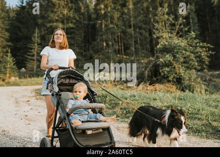 Mother with baby in stroller and dog walking on forest path Stock Photo