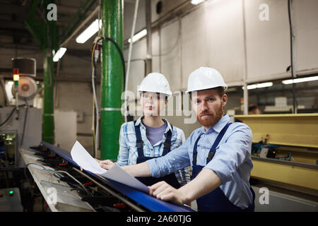 Portrait of two engineers in overalls and hardhats looking at camera while working with blueprint in the factory Stock Photo