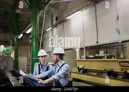 Two engineers in work helmets working over blueprint and discussing new project while standing near the lathe Stock Photo