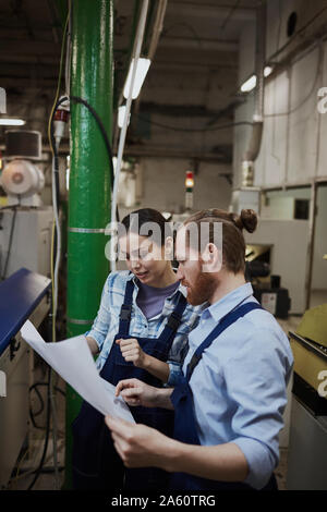 Two colleagues in overalls standing and examining blueprint together while working in the plant Stock Photo