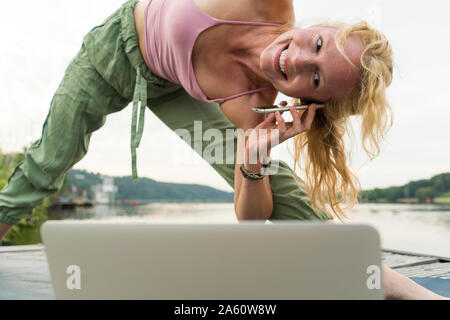Young woman stretching and using laptop and cell phone on a jetty at a lake Stock Photo