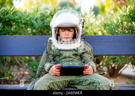 Boy wearing a space suit and sitting on a bench, playing alone with video games Stock Photo