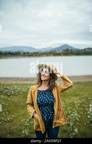 Happy young curly haired woman wearing a yellow coat and blue t-shirt holding a hat in her head with a lake in the background Stock Photo
